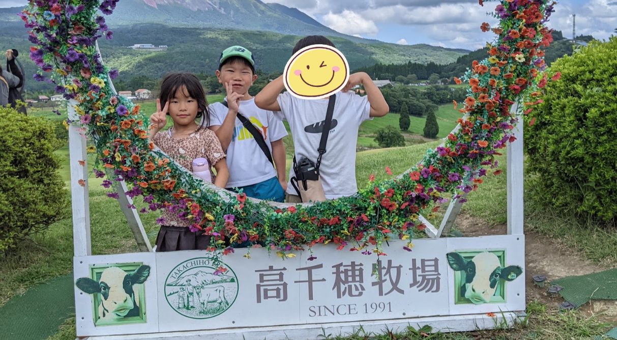 高千穂牧場の看板の前で記念撮影をする子どもたち。奥には霧島の山なみが見える